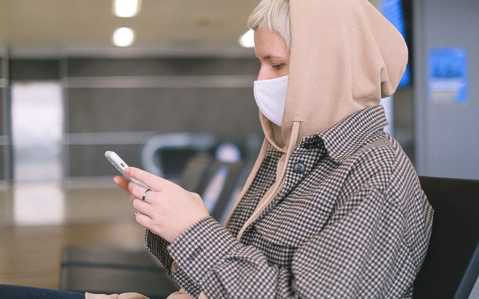 Женщина в защитной маске использует смартфон, сидя в зоне ожидания аэропорта.
